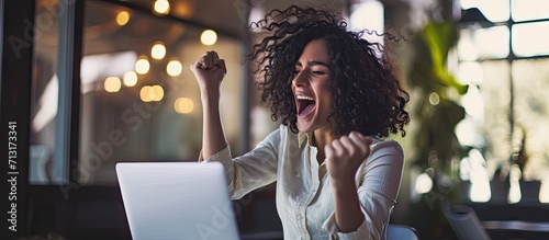 Hispanic business woman celebrating victory success employee with curly hair inside office reading good news using laptop at work inside office holding hand up and happy triumph gesture