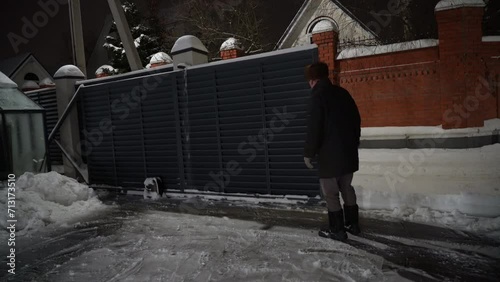 Elderly man dressed in warm clothes controls the opening of an automatic gate on a snowy winter evening. Modern designer motorized automatic gates for driveway courtyard in winter. photo