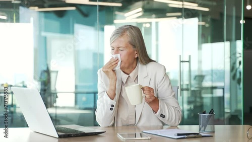 Sick senior gray haired businesswoman suffering from cold and runny nose while sitting at a workplace in a business office. Mature female wipes her nose with a handkerchief. She has a fever and flu photo