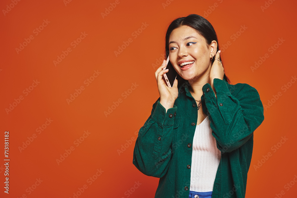 cheerful young asian woman with brunette hair talking on smartphone and smiling on orange background cheerful young asian woman with brunette hair talking on smartphone and smiling on orange background