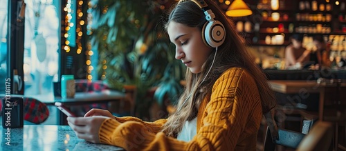 teenage woman wearing headphones sits on her phone while listening to music playing games ordering online on the sofa comfortably in a cafe. Copy space image. Place for adding text photo