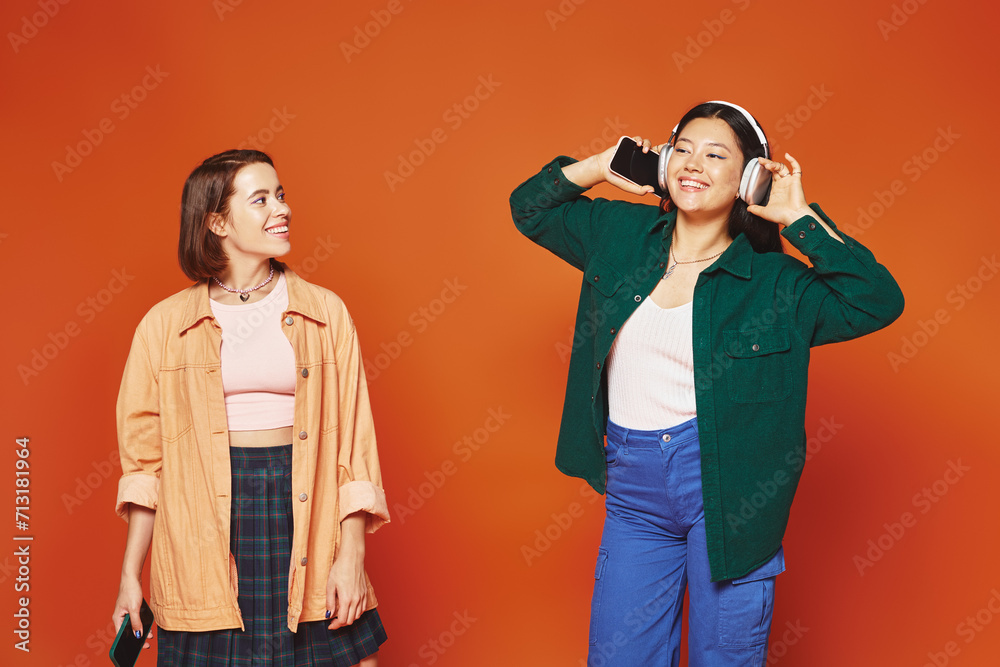 happy woman with smartphone looking at her asian friend in headphones on orange background happy woman with smartphone looking at her asian friend in headphones on orange background