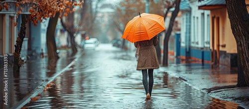 Stylish woman with orange umbrella walking on flooded street Stylish female in coat with umbrella walk on city street in rainy day Alone female with umbrella. Copy space image. Place for adding text © Ilgun