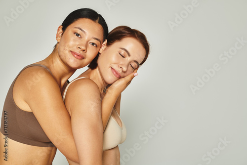 happy asian woman embracing with her female friend in underwear while posing on grey backdrop