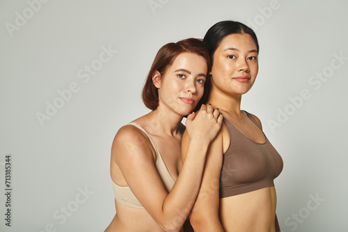 young multicultural women in underwear embracing and looking at camera on grey background