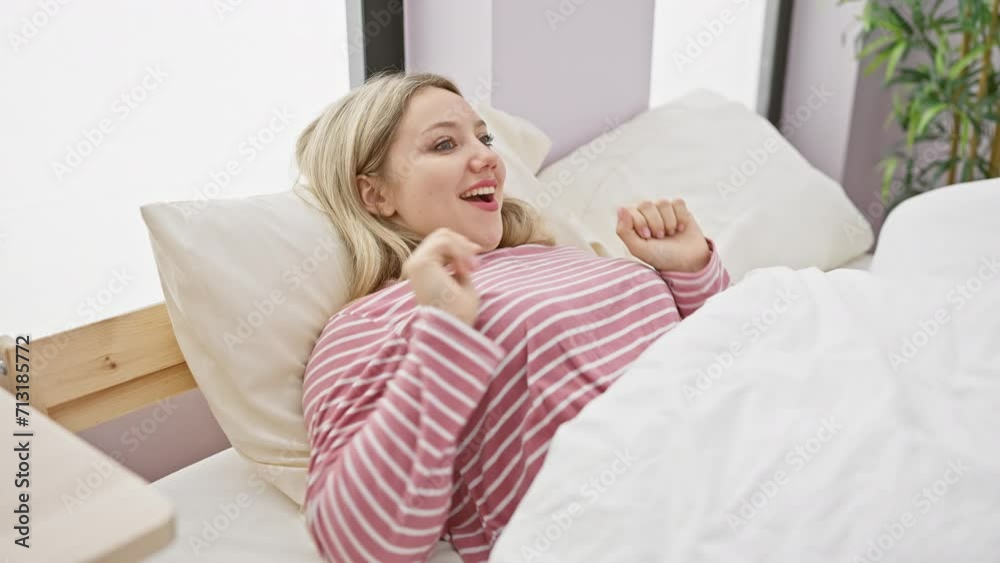 A young blonde woman in pink striped pajamas enjoys a leisurely morning in a bright bedroom, stretching and smiling with energy. A young blonde woman in pink striped pajamas enjoys a leisurely morning in a bright bedroom, stretching and smiling with energy.