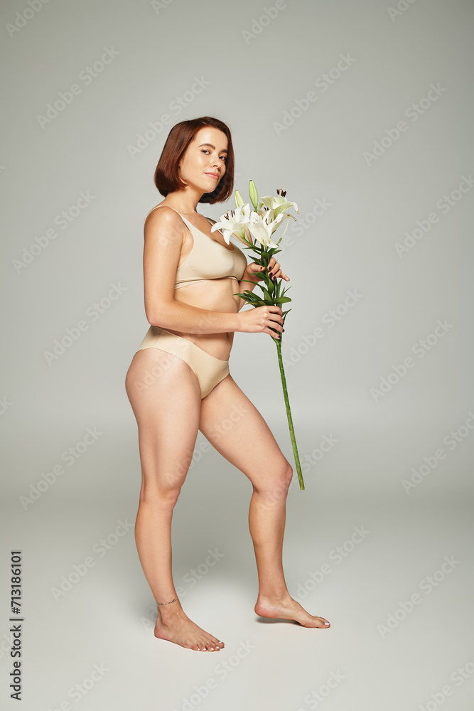 full length of woman with short hair holding flowers and posing in underwear on grey background
