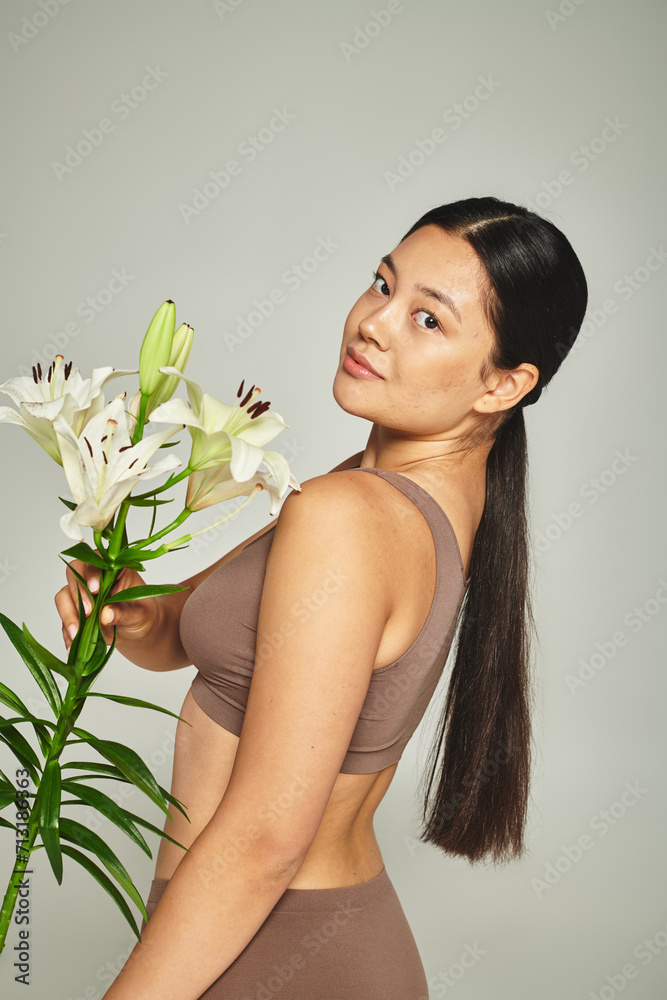 brunette asian woman in pastel underwear holding flowers and standing on grey background, lilies brunette asian woman in pastel underwear holding flowers and standing on grey background, lilies