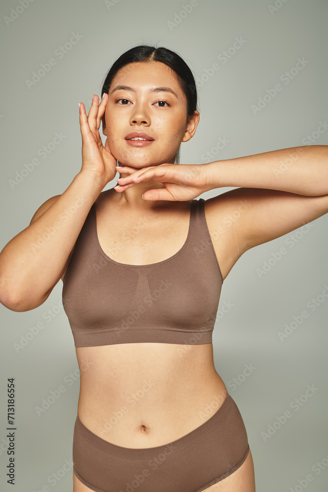 brunette asian woman in brown top touching her face on grey background, skin care and beauty brunette asian woman in brown top touching her face on grey background, skin care and beauty