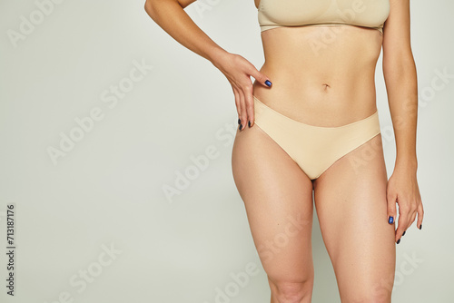 cropped shot of young woman standing in beige lingerie with hand on hip on grey background