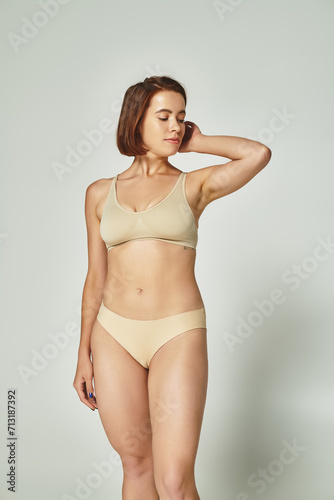 body positive, pretty young woman with short hair standing in beige underwear on grey background