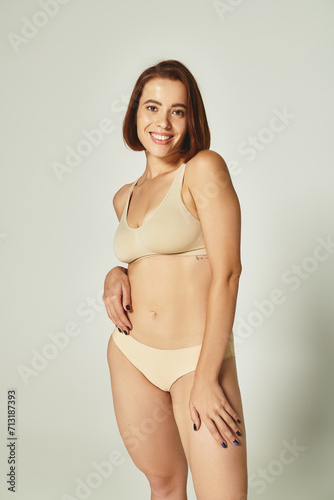 body positive, cheerful young woman with short hair standing in beige underwear on grey background © LIGHTFIELD STUDIOS