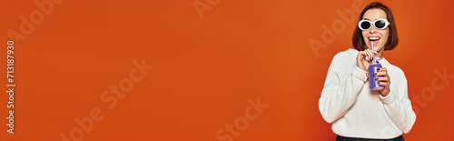 Quadro em tela Stylish young woman in white trendy sunglasses sipping soda drink on orange back