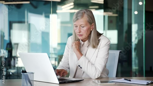 Serious thoughtful elderly senior gray haired businesswoman works on a laptop sitting at a desk at a workplace in a modern office. Mature business female is busy with project on computer, thinking photo