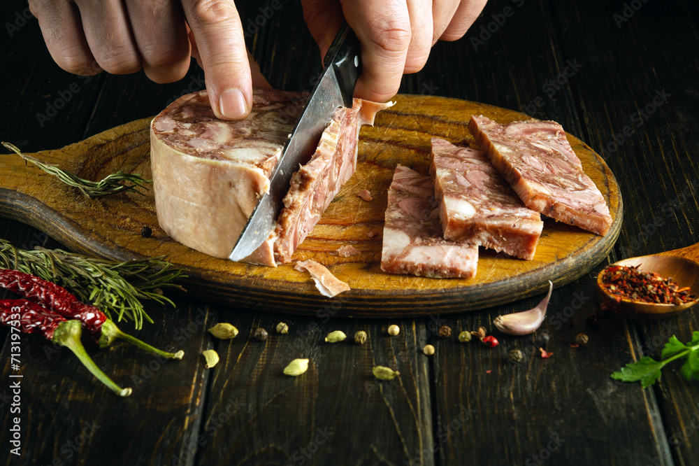 The cook is preparing a meat dish for lunch. Slicing meat brawn with a knife in the hand of a chef on a kitchen board