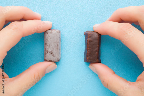 Young adult woman hand fingers holding fresh and old spoiled dark chocolate candies on light blue table background. Pastel color. Compare two pralines. Point of view shot. Top down view.