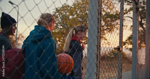 A happy blonde girl together with her friends with a basketball, enters a special basketball court fenced with a grid for playing basketball early in the morning at sunrise photo