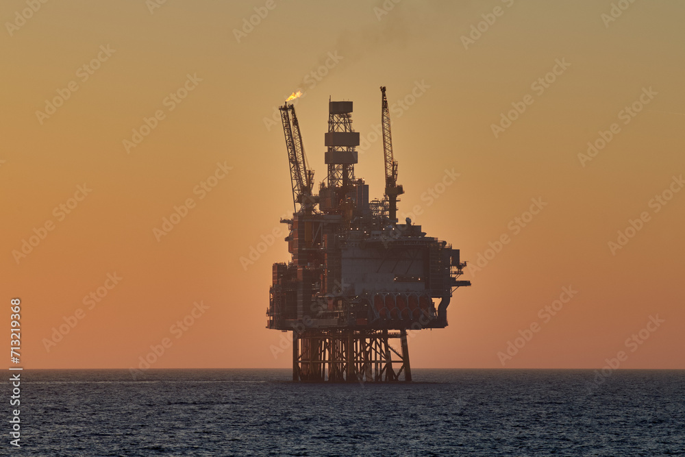 Silhouette of offshore oil and gas jackup platform in the ocean during sunset, with yellow sky and calm water.