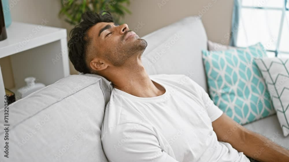 A relaxed hispanic man lounges in a cozy living room on a gray sofa with decorative pillows, embodying casual home comfort.