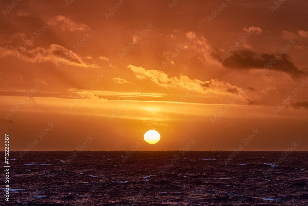 Orange ocean sunset, with clouds, blue ocean and sun on the horizon.