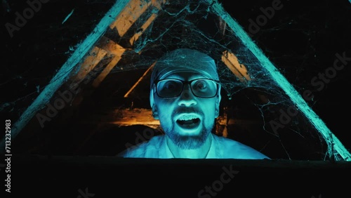 A mad scientist with glasses lit by a blue glow grimaces and screams looking at the camera, surrounded by cobwebs and wooden beams photo