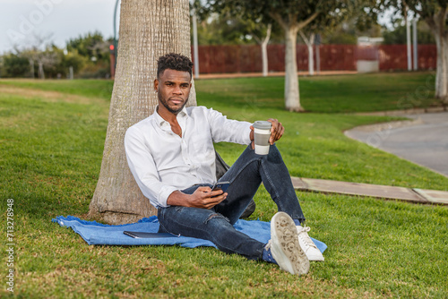 A man sits comfortably in the green grass, leaning against a tall tree, enjoying the warmth of a sunny day while holding a cell phone and a cup of coffee. photo