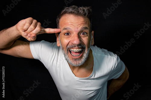 Portrait of a mature man with a gray beard, wearing braces, gesturing with his hands, wearing white t-shirt on a black background.