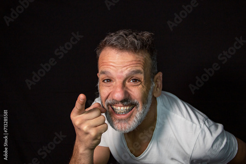 Portrait of a mature man with a gray beard, wearing braces, gesturing with his hands, wearing white t-shirt on a black background.