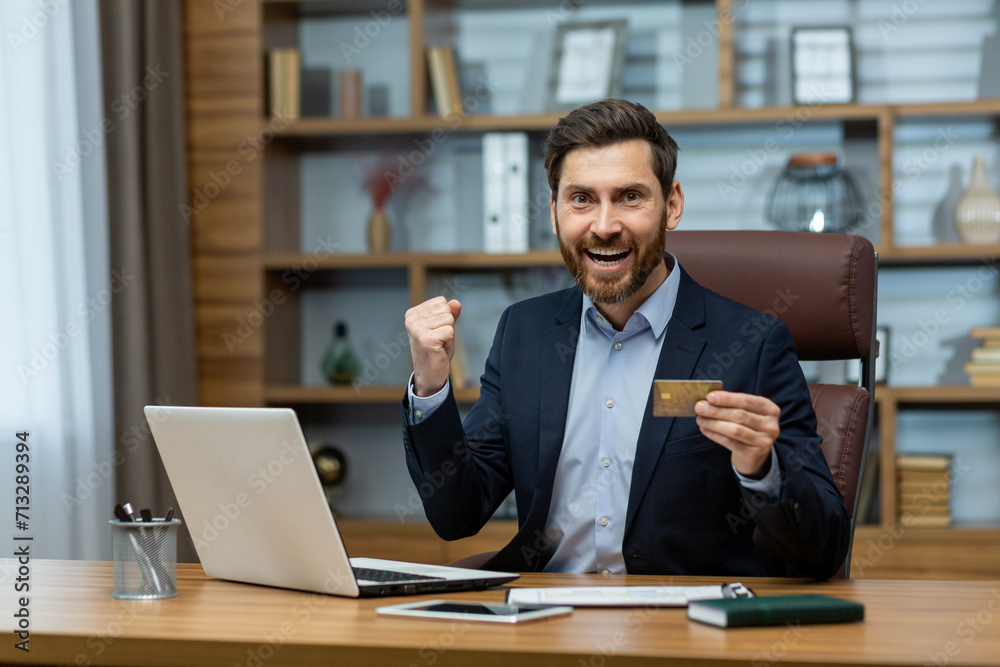Obraz premium Joyful mature businessman holding a credit card and celebrating a financial triumph in a tastefully decorated home office.