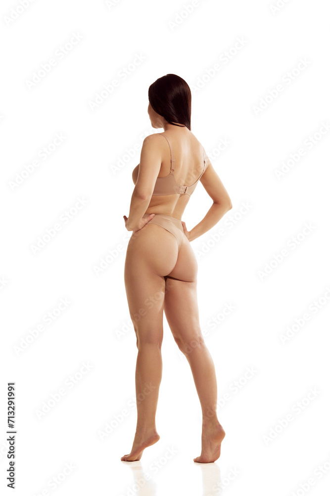 Naklejka premium Back view portrait of young woman with perfect curves, without cellulite posing put her hands on hips against white studio background. Concept of health, beauty, youth, spa, fitness, ad