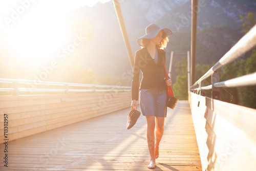 Elegant Caucasian Adult Woman Walking Barefoot over a Modern Wooden Suspended Bridge Backlit from Sunset Warm Light in Mountains - Lago Di Cavazzo, Udine, Italy photo