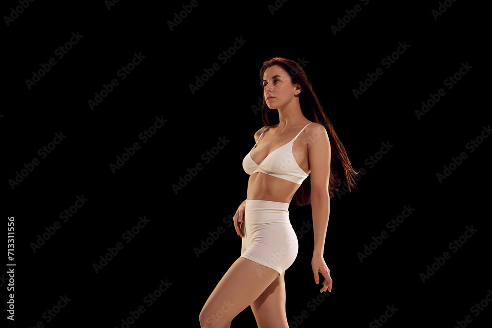 Side view portrait of beautiful fit young woman in white sportwear posing with arms up against dark mode background. Concept of female health, fitness, sport, active lifestyle, dieting.