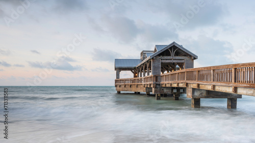 Wooden pier, with a covered open air structure at the end, reaching out into the tropical waters of the ocean, at sunrise. There is a hint of orange in the sky