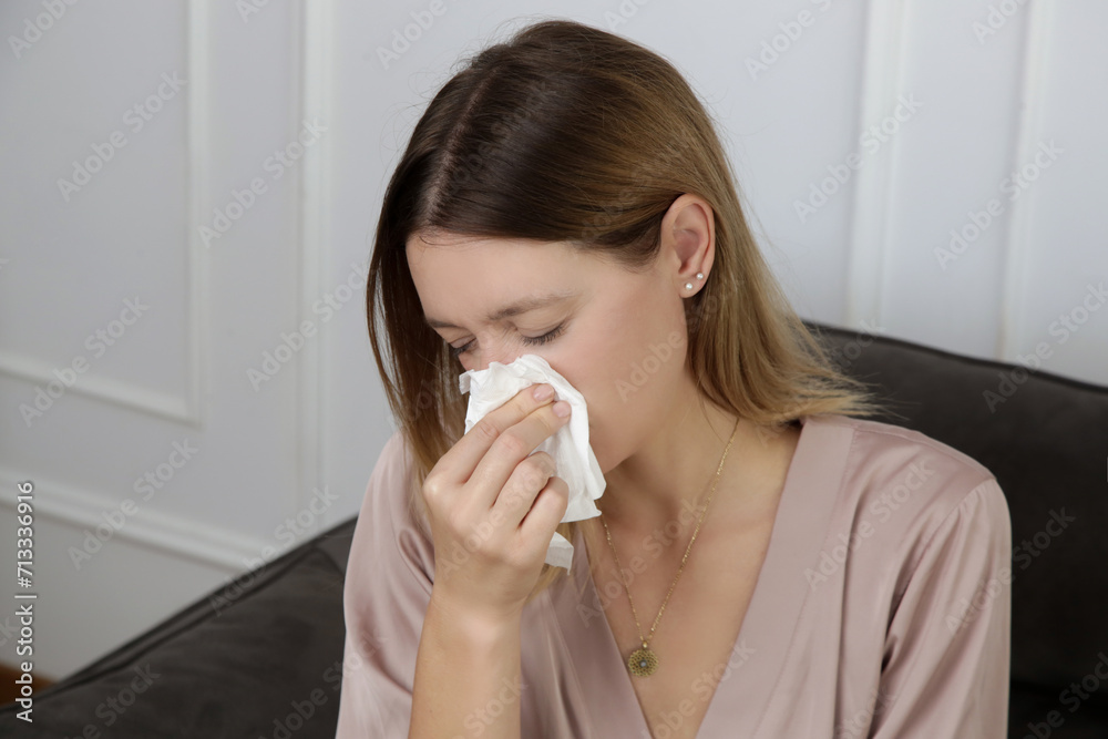 Young woman on the sofa blowing nose into a white paper tissue. Girl with allergy symptoms sneezing into a tissue. Flu, cold or allergy symptom	
