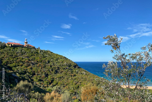 Red lighthouse at the top of a green mountain. Hill, beach and bay in the ocean. View of the coast of island. Luxurious landscape. Sydney, Australia.