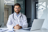 Portrait of a smiling young male doctor in a white coat sitting in the office of the clinic at the working table and confidently looking at the camera