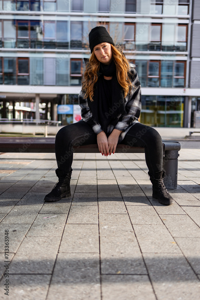 Woman in her mid-thirties with long red hair wearing a cap and black clothing in an urban location.