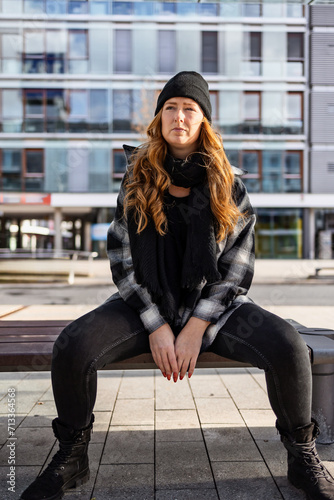 Woman in her mid-thirties with long red hair wearing a cap and black clothing in an urban location.