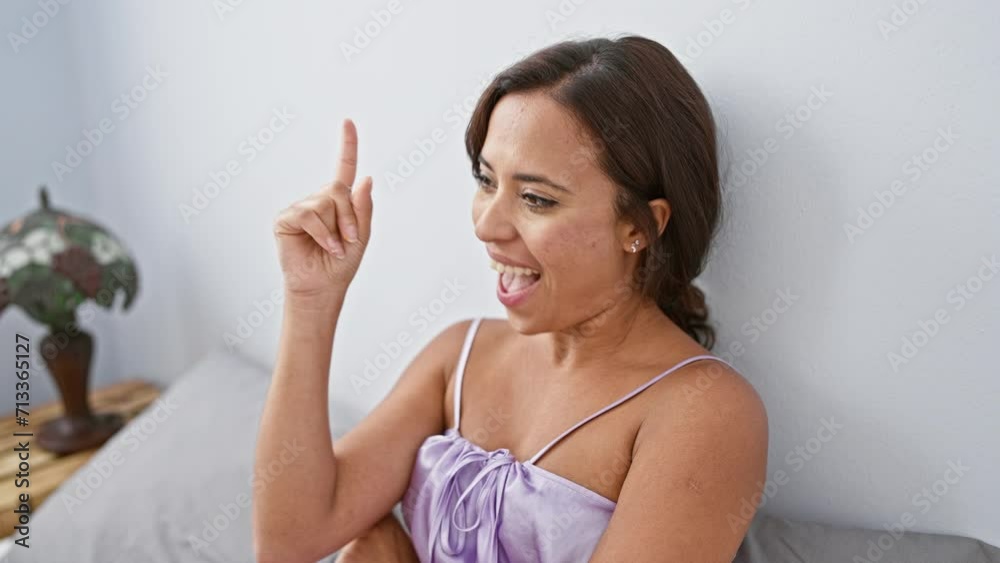 Cheerful young hispanic woman in bedroom, lounging in bed wearing pyjama. admire her as she smiles, cleverly gesturing and pointing finger up, signifying number one success idea! feel-good face. Cheerful young hispanic woman in bedroom, lounging in bed wearing pyjama. admire her as she smiles, cleverly gesturing and pointing finger up, signifying number one success idea! feel-good face.
