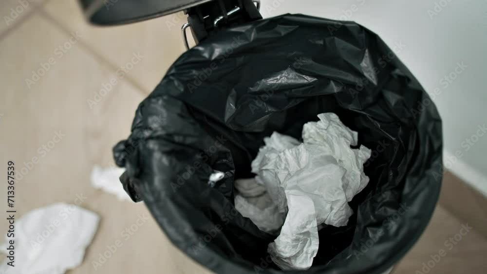 A person throws a napkin into a bucket with a pedal to open it. white paper nets fall over a metal trash can and past it onto the floor. A man opens garbage and throws used paper napkins for recycling A person throws a napkin into a bucket with a pedal to open it. white paper nets fall over a metal trash can and past it onto the floor. A man opens garbage and throws used paper napkins for recycling