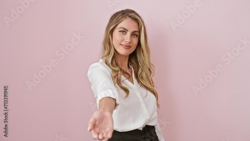 Cheerful young blonde woman in shirt, standing over isolated pink background, offering a friendly handshake gesture of acceptance and assistance, radiating positivity and heartwarming welcome