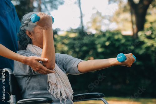 Young nurse or physiotherapist in scrubs helping a happy retired old woman do fitness exercises with light weight dumbbells at home. Concept of physiotherapy for seniors