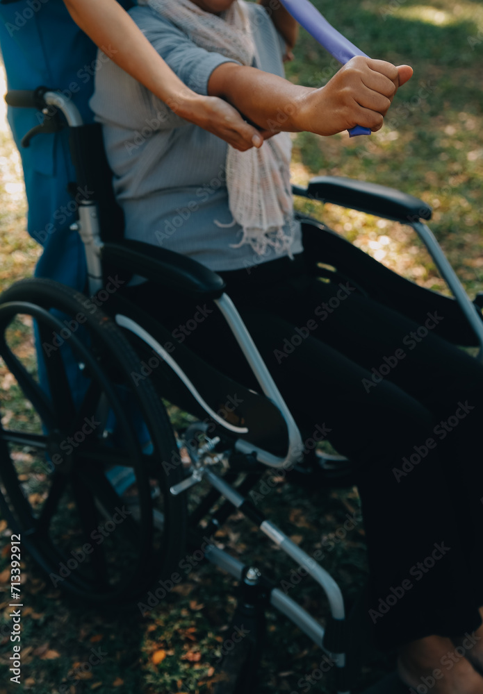 Naklejka premium Young nurse or physiotherapist in scrubs helping a happy retired old woman do fitness exercises with light weight dumbbells at home. Concept of physiotherapy for seniors