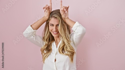 Playful young woman posing, blonde in shirt making fun bull horns with fingers over head, on isolated pink background photo