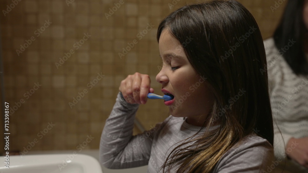 Daughter brushing teeth in front of bathroom mirror by sink while mother helps smaller brother to brush teeth, authentic family dental hygiene Daughter brushing teeth in front of bathroom mirror by sink while mother helps smaller brother to brush teeth, authentic family dental hygiene