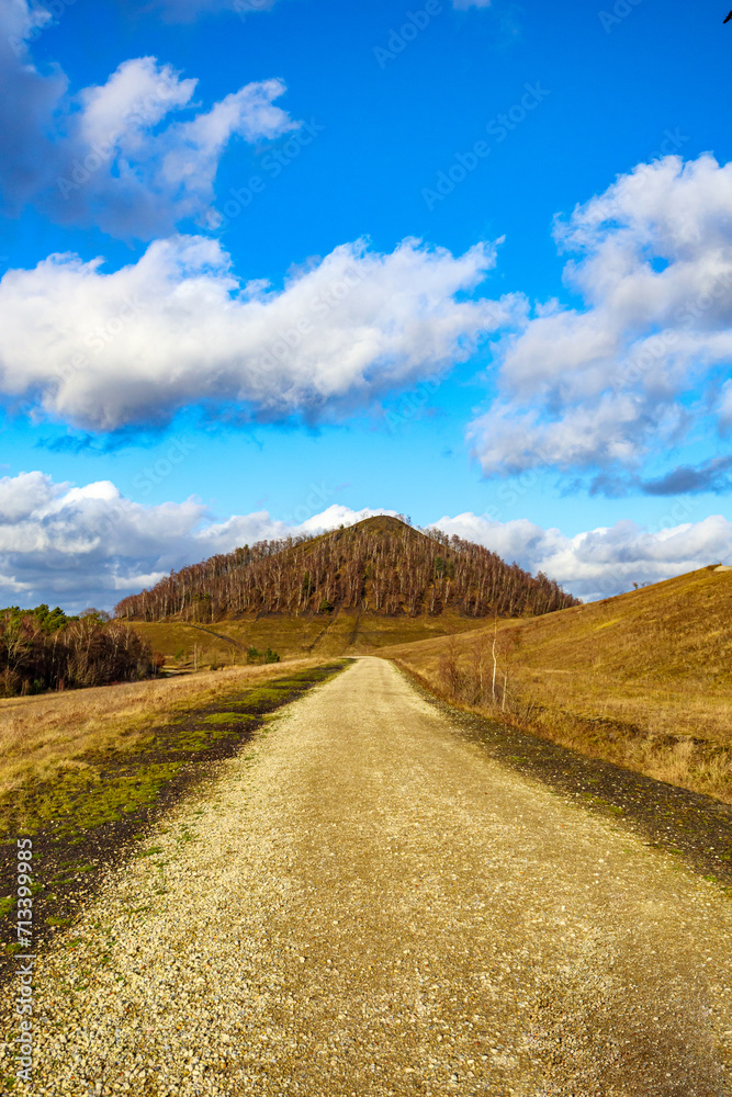 Front view of dirt road between plains and hills, mountain with autumn trees against blue sky with white clouds in background, Thor Park - Hoge Kempen National Park, sunny afternoon in Genk, Belgium