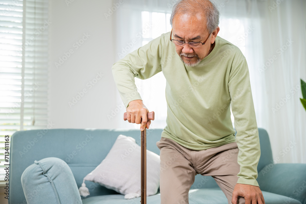 Elderly man suffering from knee pain ache holding handle of cane, senior old man typing to stand up from sofa alone with walking cane stick to walk at home, retirement medical healthcare concept Elderly man suffering from knee pain ache holding handle of cane, senior old man typing to stand up from sofa alone with walking cane stick to walk at home, retirement medical healthcare concept