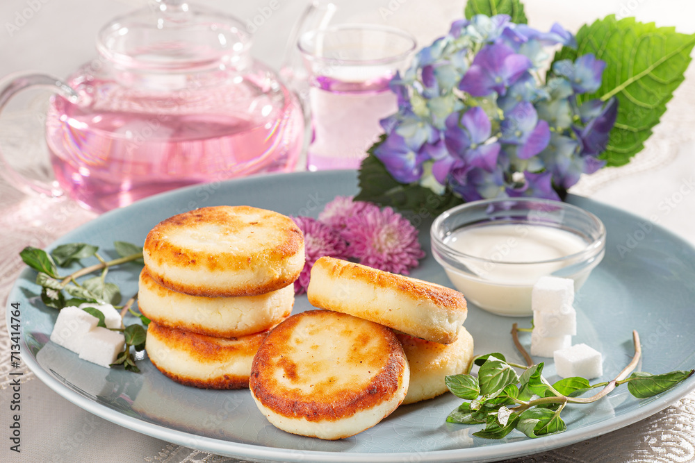 Fried cottage cheese pancakes syrniki served with sour cream and flower tea on a blue dish.