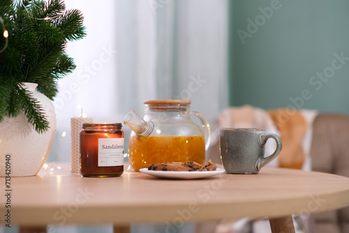 Home kitchen interior- glass hot teapot with sea buckthorn tea , plate with biscuits, burning candle, vase with fir branches on wooden table near the window.