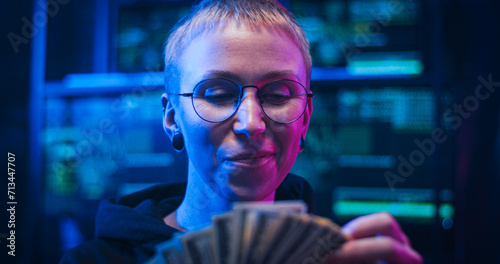 Close up of Caucasian female hacker in glasses counting money in dark room with many monitors. Woman cyber criminal holding dollars banknotes in hands. Dirty salary and laundering concept.